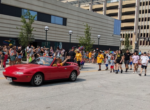People waving from Red Miata in parade. Large speakers sitting in back seat.
