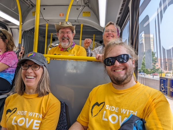 Pride Parade goers sitting in ORBT bus, wearing yellow Side with Love t-shirts.