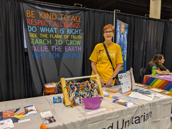 Woman standing in Second Unitarian Church's Pride booth. Banner hanging from back of booth displays the words, "Be kind to all; Respect all beings; Do what is right; Seek the flame of truth; Search to grow; Value the earth; and Fight for justice."