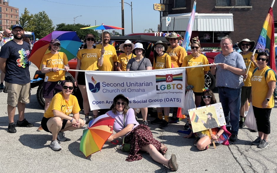 Twenty people of all ages are gathered just prior to marching in the 2024 Heartland Pride Parade. Two are holding a banner that bears the words, "Second Unitarian Church of Omaha" and "Open Arms Trans Social Group (OATS)" with a "LGBTQ" rainbow logo. Many of the people are wearing yellow shirts with "Side With Love" printed on them. Several are holding rainbow-striped umbrellas. A young girl is holding a poster she made for the parade.